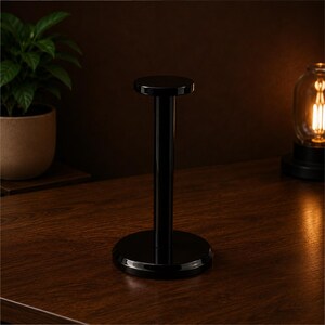 May include: A glossy black headphone stand featuring a circular base, a vertical cylindrical support column, and a flat circular top platform. The stand has a smooth, reflective finish and a simple, functional design for displaying headphones.
