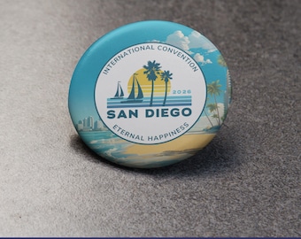 Puede incluir: Un botón redondo con un diseño azul y blanco que promociona la Convención Internacional 2026 en San Diego. El botón presenta una escena de playa con palmeras, veleros y el texto "San Diego Eternal Happiness."