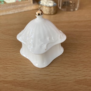 May include: A white porcelain trinket box with a gold-coloured knob on the lid. The box has a scalloped edge and a decorative, embossed design. The box is on a wooden surface.
