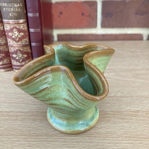 May include: A small, handcrafted ceramic bowl with a unique, ruffled rim. The bowl features a light green glaze with brown accents, and a pedestal base. The bowl is displayed on a wooden surface, with vintage books in the background.