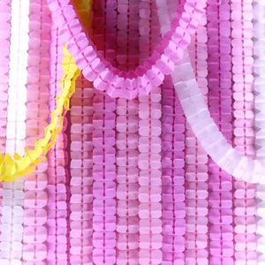 May include: A close-up of colourful paper garlands in shades of pink, white, and yellow. The garlands are made of folded paper and are arranged vertically and horizontally, creating a textured background. The paper garlands are suitable for party decorations.