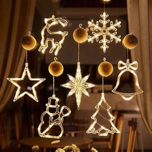 Può includere: Una collezione di decorazioni natalizie illuminate, tra cui una renna, una stella, un fiocco di neve, un pupazzo di neve, una stella, una campana e un albero di Natale. Le luci bianche calde sono appese a una finestra.