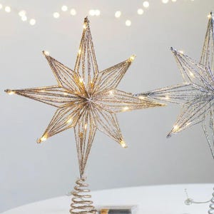 May include: Glittering star-shaped Christmas tree toppers with warm white LED lights. The gold star has a spiral base and is next to a silver star. These decorative ornaments add a festive touch to holiday decor.