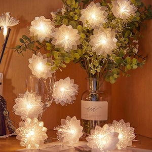 Pode incluir: Uma sequência de luzes decorativas em forma de flores brancas. As luzes estão dispostas em torno de um vaso de vegetação e um copo de vinho. As luzes estão acesas, criando um brilho quente. As flores têm uma textura brilhante.