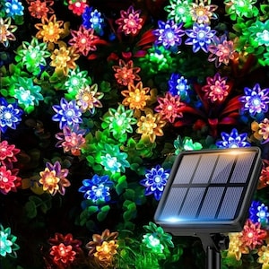 May include: Solar-powered string lights with flower-shaped LEDs in a variety of colours, including red, blue, green, and yellow. The lights are set amongst green foliage, with a black solar panel in the foreground.
