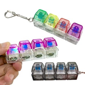 May include: A selection of colourful mechanical keyboard keycaps. The keycaps are clear with various coloured tops, including green, yellow, red, and purple. Some keycaps are attached to a keychain. The keycaps are lit with blue lights.