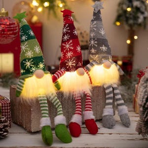 May include: Three Christmas gnome decorations with illuminated fluffy white beards and striped legs. The gnomes have tall, pointed hats in green, red, and gray, each adorned with snowflake patterns. The gnomes are sitting on a wooden block.