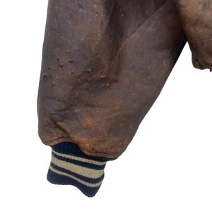 May include: Close-up of a distressed brown leather jacket sleeve. The sleeve shows wear and tear, with a ribbed cuff featuring navy blue and beige stripes. The leather has a worn, vintage appearance.