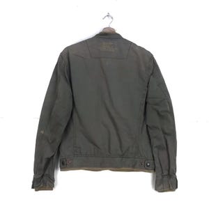 Puede incluir: Chaqueta verde oliva con cierre de botones, cuello y mangas largas. La parte trasera de la chaqueta tiene un peque&ntilde;o parche con texto. La chaqueta cuelga de una percha met&aacute;lica sobre un fondo blanco.