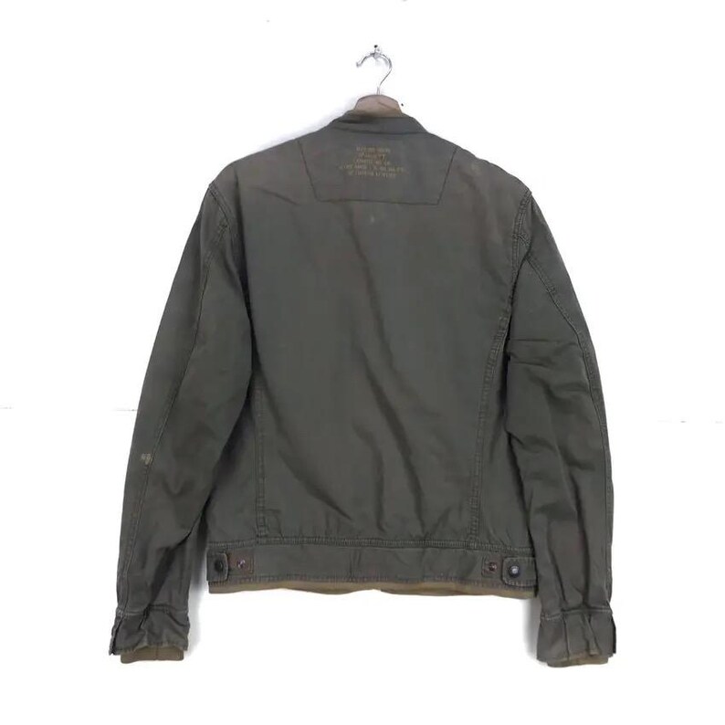 Puede incluir: Chaqueta verde oliva con cierre de botones, cuello y mangas largas. La parte trasera de la chaqueta tiene un peque&ntilde;o parche con texto. La chaqueta cuelga de una percha met&aacute;lica sobre un fondo blanco.