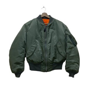 May include: An olive green bomber jacket with a black zip, pockets, and ribbed cuffs. The jacket has a dark grey collar and an orange interior lining. The jacket is hanging from a wooden hanger.