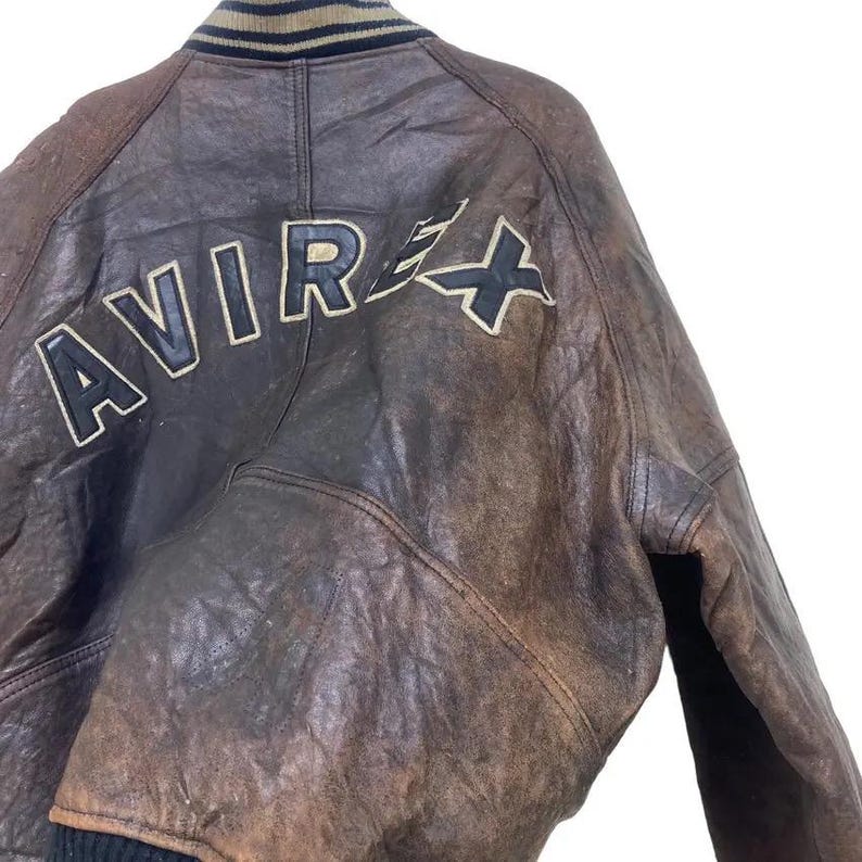 May include: Brown leather jacket with the word "AVIREX" in white letters on the back. The jacket has a black and tan striped collar and cuffs. The leather has a distressed appearance.