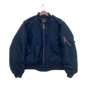 May include: A navy blue bomber jacket with a ribbed collar, cuffs, and hem. The jacket features a zipper closure, two front pockets, and a small pocket on the left sleeve. The jacket has a dark blue patch on the front and brown leather zipper pulls.