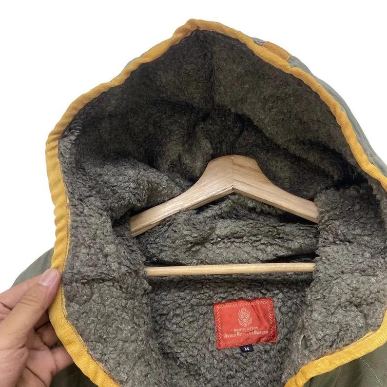 Op de afbeelding: Close-up van een olijfgroene jas met capuchon en gele rand. De binnenkant is gevoerd met een grijs, fuzzy materiaal. Een rood rechthoekig label met witte tekst is zichtbaar. De jas hangt aan een houten hanger.