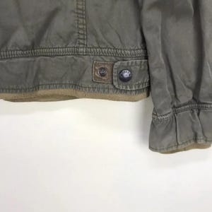 Puede incluir: Primer plano de una chaqueta verde oliva con un parche de cuero marr&oacute;n y un bot&oacute;n. La chaqueta tiene un dobladillo y pu&ntilde;os acanalados. La chaqueta est&aacute; hecha de un material duradero.