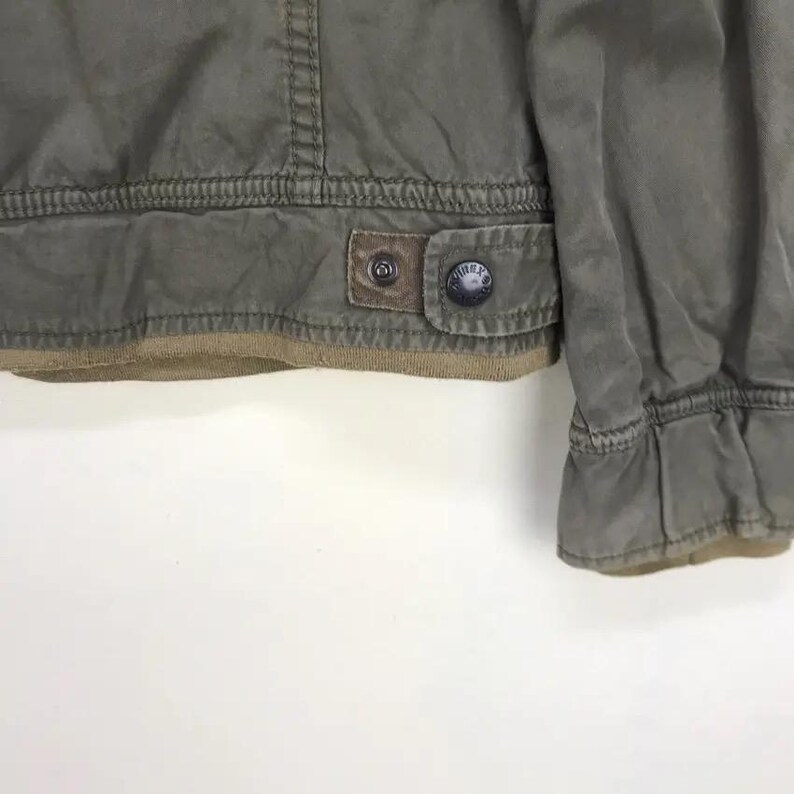 Puede incluir: Primer plano de una chaqueta verde oliva con un parche de cuero marr&oacute;n y un bot&oacute;n. La chaqueta tiene un dobladillo y pu&ntilde;os acanalados. La chaqueta est&aacute; hecha de un material duradero.