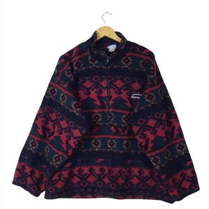 Puede incluir: Un jersey polar con un estampado geométrico en rojo, azul marino y verde. La chaqueta tiene cuello alto, media cremallera y un pequeño logotipo en el pecho. El diseño presenta rombos y formas angulares.