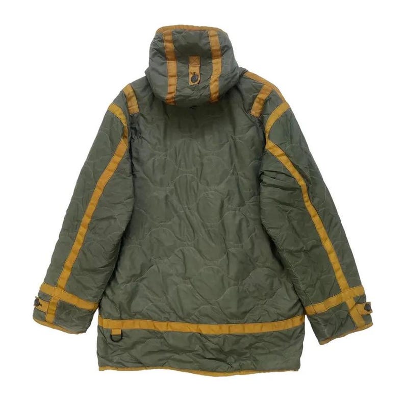 Op de afbeelding: Olijfgroene gewatteerde jas met capuchon en goudkleurige accenten. De jas heeft gouden strepen langs de mouwen en rond de onderkant. De jas heeft een gewatteerd patroon en een D-ring detail.