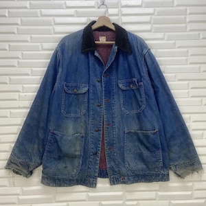 May include: A vintage denim jacket with a dark blue collar and multiple pockets. The jacket has a button-up front and a red interior lining. The denim is faded, giving it a worn look. The jacket is hanging on a metal hook.