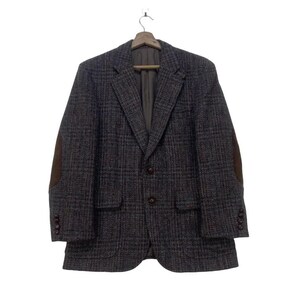 May include: A dark gray tweed blazer with brown elbow patches, two front pockets, and brown buttons. The blazer is hanging on a wooden hanger against a white background. The jacket has a classic, tailored design.
