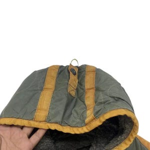 Op de afbeelding: Close-up van een groene gewatteerde jaskap met bruine accenten. De capuchon heeft een knoopsluiting en een zachte, getextureerde voering. Het ontwerp van de jas omvat verticale bruine strepen en een kleine lus.