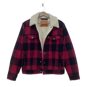 May include: Red and black plaid jacket with a cream-colored sherpa collar and lining. The jacket has two chest pockets and button closures. A small Levi's tag is visible on the chest. The jacket is hanging on a wooden hanger.