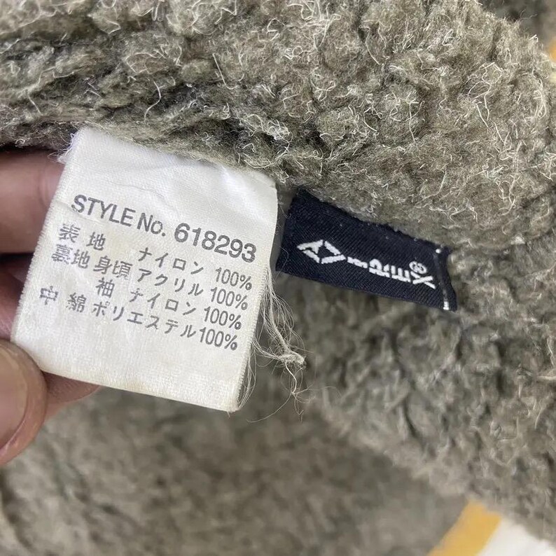 Op de afbeelding: Close-up van een getextureerde, olijfgroene stof met een wit label en een zwart label. Het witte label toont de tekst "STYLE No. 618293" samen met Japanse tekst die de stofsamenstelling aangeeft. Het zwarte label heeft een wit logo.