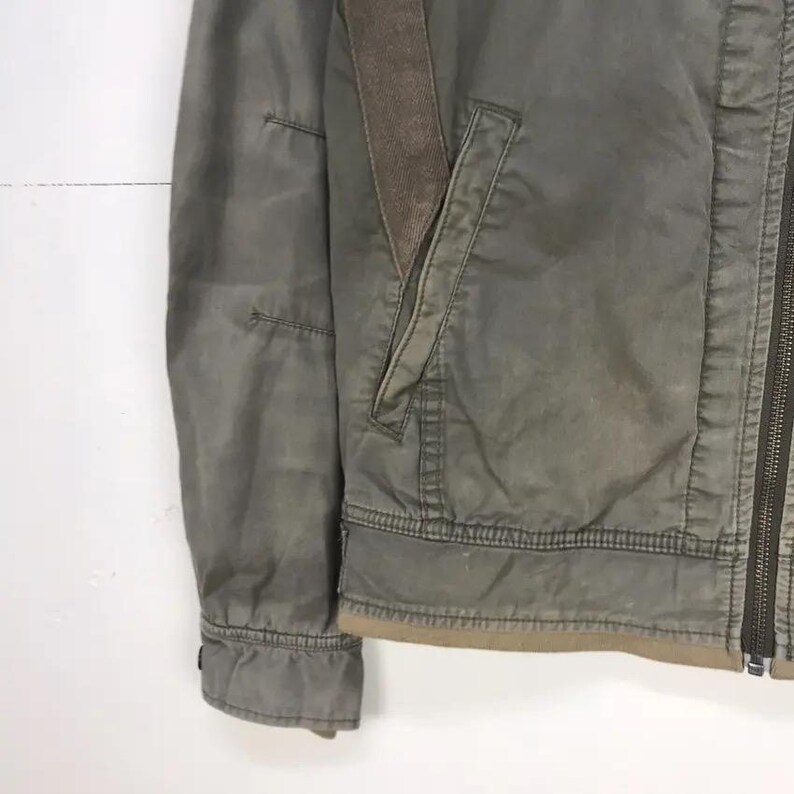 Puede incluir: Primer plano de una chaqueta verde oliva desgastada con un acento marr&oacute;n en el hombro y el bolsillo. La chaqueta tiene cierre de cremallera y un ribete marr&oacute;n en la parte inferior. La tela parece ser un material duradero, posiblemente lona.