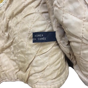 May include: Close-up of a quilted, beige fabric with a diamond pattern. A navy blue tag is sewn into the fabric, displaying the text "KOREA EN COR&Eacute;E" in white lettering. The fabric appears to be part of a garment lining.