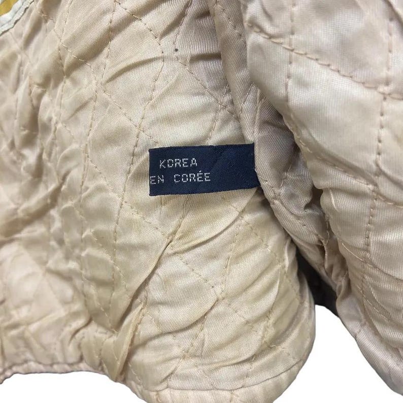 May include: Close-up of a quilted, beige fabric with a diamond pattern. A navy blue tag is sewn into the fabric, displaying the text "KOREA EN COR&Eacute;E" in white lettering. The fabric appears to be part of a garment lining.