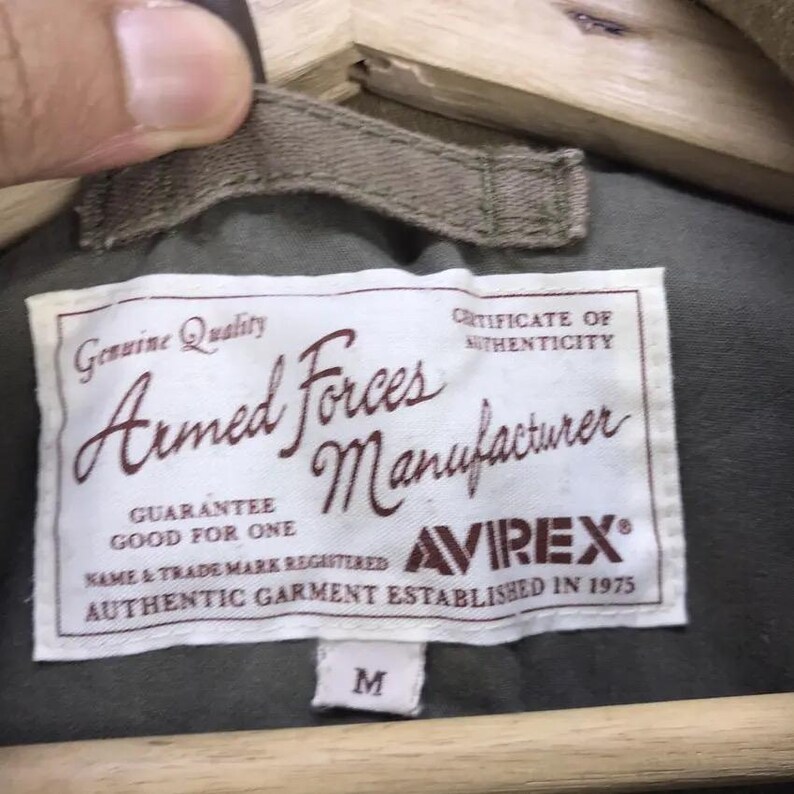 Puede incluir: Primer plano de una etiqueta blanca con el texto "Armed Forces Manufacturer" en un estilo vintage. La etiqueta tambi&eacute;n incluye la marca "AVIREX" y el texto "Authentic Garment Established in 1975". La etiqueta est&aacute; cosida a una prenda verde.
