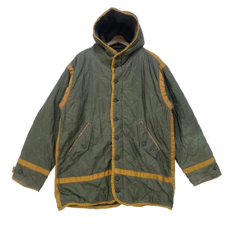 Op de afbeelding: Olijfgroene gewatteerde jas met capuchon en knoopsluiting. De jas heeft gele biezen langs de randen en zakken. De jas heeft een vintage uitstraling.