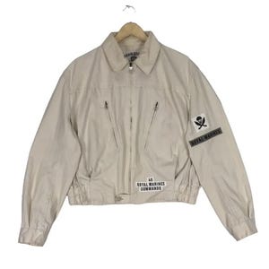 May include: A beige jacket with a collar, zipper closure, and multiple pockets. The jacket features a "Royal Marines" patch with a skull and crossbones emblem and the text "45 Royal Marines Commando".