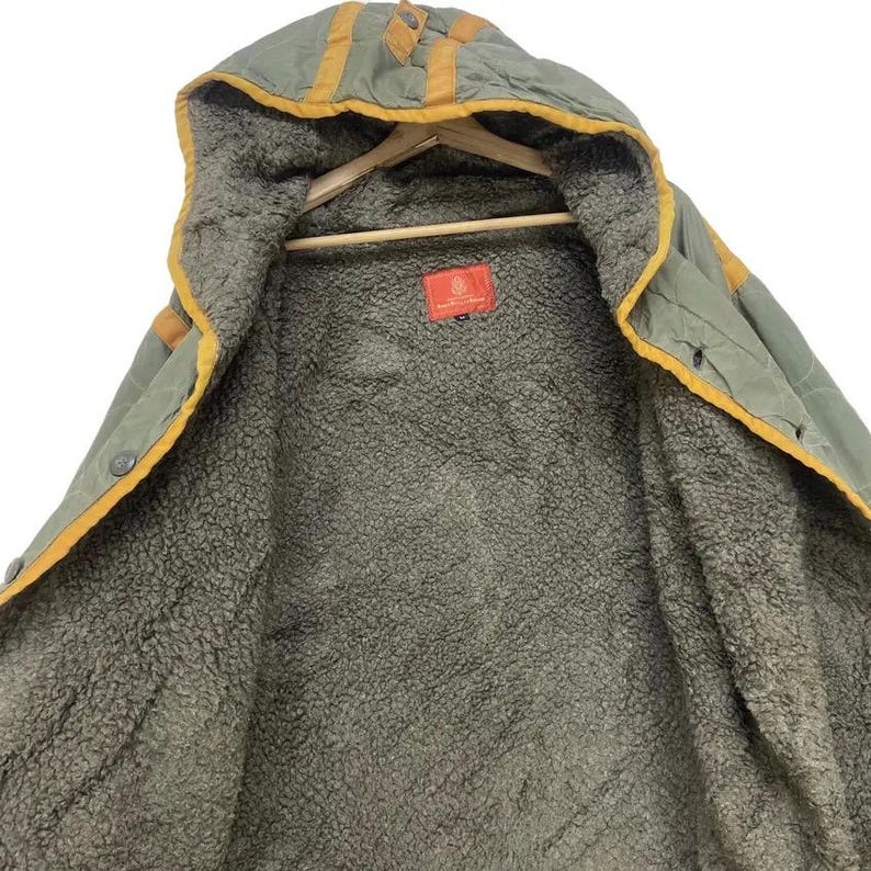 Op de afbeelding: Olijfgroene jas met capuchon en een zachte, fuzzy binnenkant. De jas heeft gele biezen langs de capuchon en naden, met een knoopsluiting. Een rood label is zichtbaar aan de binnenkant.