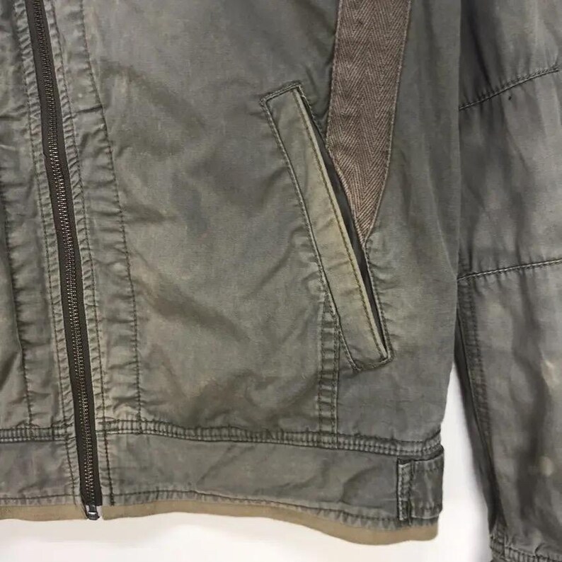 Puede incluir: Primer plano de una chaqueta verde con cremallera completa y detalle de espiga marr&oacute;n en el hombro. La chaqueta tiene un bolsillo en el pecho y un aspecto desgastado.