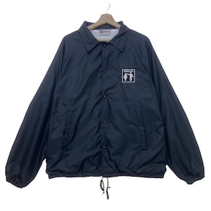 以下が含まれることがあります： ボタン留め、襟、ドローコードの裾が付いた黒いコーチジャケット。胸には「bitch」の文字と2人の人物のグラフィックが入った白いロゴが付いています。ジャケットは滑らかな、おそらくナイロン素材で作られています。