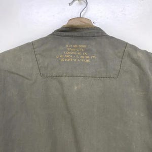 Puede incluir: Una chaqueta verde oliva descolorida cuelga de una percha met&aacute;lica. La parte trasera de la chaqueta tiene texto dorado, con medidas: 116.8 cm de largo y un peso de 1.5 kg. El cuello es de color marr&oacute;n claro.