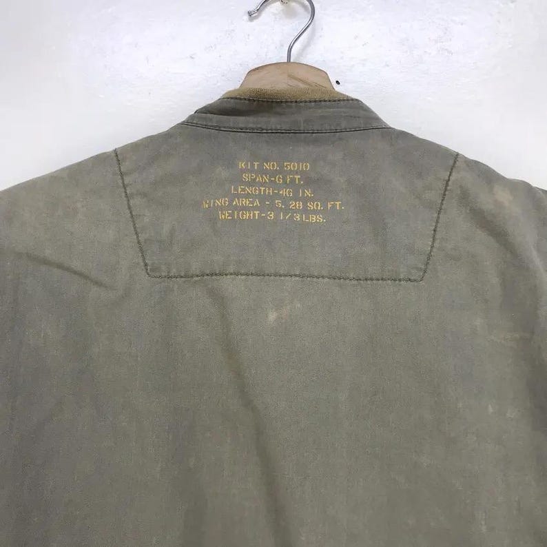 Puede incluir: Una chaqueta verde oliva descolorida cuelga de una percha met&aacute;lica. La parte trasera de la chaqueta tiene texto dorado, con medidas: 116.8 cm de largo y un peso de 1.5 kg. El cuello es de color marr&oacute;n claro.