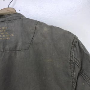 Puede incluir: Primer plano de una chaqueta verde oliva descolorida. La chaqueta tiene costuras visibles a lo largo de las costuras y un cuello. Texto tenue visible en la parte superior izquierda. La chaqueta est&aacute; colgada de una percha de madera.