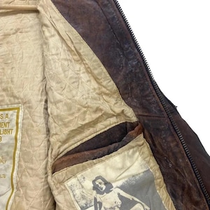 May include: Close-up of a brown leather jacket interior. The jacket has a quilted beige lining and a pocket with a vintage graphic. The jacket features a zipper and a label with text. The leather has a distressed appearance.