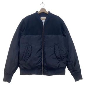 Puede incluir: Chaqueta bomber negra con un panel superior negro en contraste. La chaqueta presenta una cremallera frontal, dos bolsillos delanteros y puños y dobladillo de canalé. La chaqueta está colgada de una percha de madera.