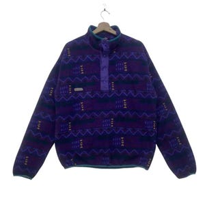 May include: A purple fleece pullover with a geometric pattern in dark blue, green, and yellow. The long-sleeved top has a button-up collar with a teal trim. A small rectangular patch is on the left chest.
