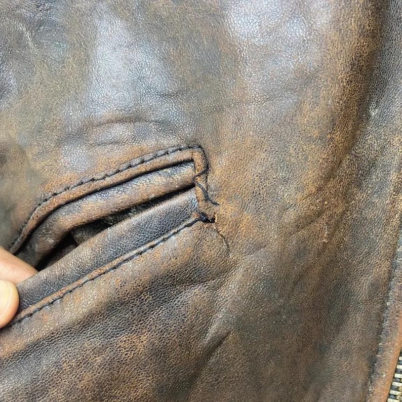 May include: Close-up of a distressed brown leather jacket, highlighting the texture and stitching. The jacket has a worn, vintage look with visible wear and tear, including a pocket and zipper.