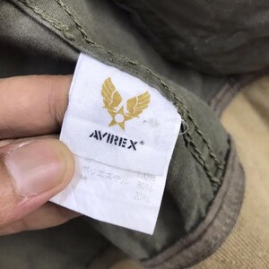 Puede incluir: Primer plano de una etiqueta blanca con el nombre de la marca AVIREX y un emblema dorado de alas y una estrella. La etiqueta est&aacute; adherida a una tela verde oliva. La etiqueta tambi&eacute;n tiene texto japon&eacute;s.