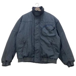 Peut inclure: Veste de bombardier gris foncé avec un col montant et plusieurs poches. La veste a une fermeture éclair et des poignets et un ourlet côtelés. Le tissu semble être lisse, possiblement résistant à l'eau, adapté aux températures plus fraîches.