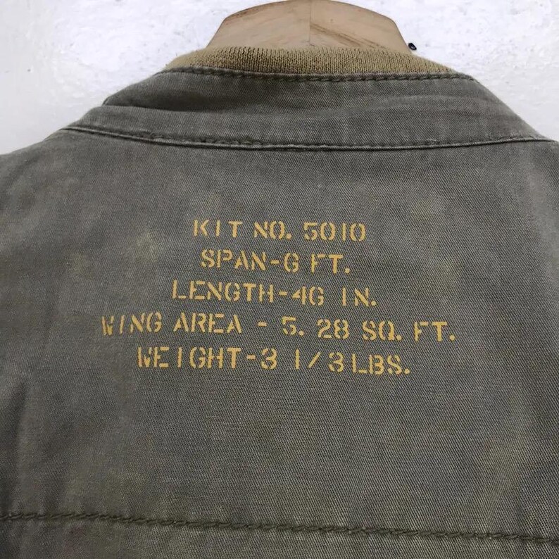 Puede incluir: Primer plano de una chaqueta verde oliva descolorida con cuello beige. Texto dorado: KIT NO. 5010, ENVERGADURA - 1,83 m, LONGITUD - 116,8 cm, SUPERFICIE ALAR - 0,49 m&sup2;, PESO - 1,5 kg. La chaqueta es de tela resistente.