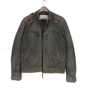 Puede incluir: Chaqueta verde oliva con parches marrones en los hombros y cremallera. La chaqueta tiene dos bolsillos delanteros y un peque&ntilde;o parche en el pecho con texto. La chaqueta est&aacute; colgada en una percha de madera.