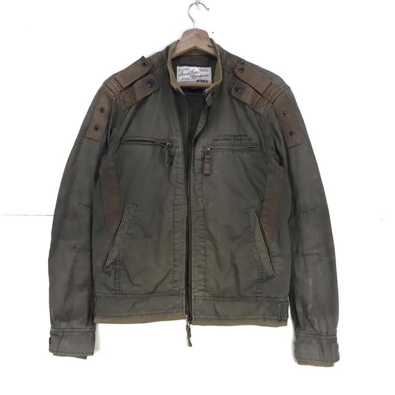 Puede incluir: Chaqueta verde oliva con parches marrones en los hombros y cremallera. La chaqueta tiene dos bolsillos delanteros y un peque&ntilde;o parche en el pecho con texto. La chaqueta est&aacute; colgada en una percha de madera.