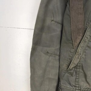 Puede incluir: Primer plano de una chaqueta verde oliva con un detalle de patr&oacute;n de espiga marr&oacute;n en el hombro. La chaqueta tiene un bolsillo y detalles de costura visibles. La tela parece ser un material duradero, posiblemente resistente al agua.