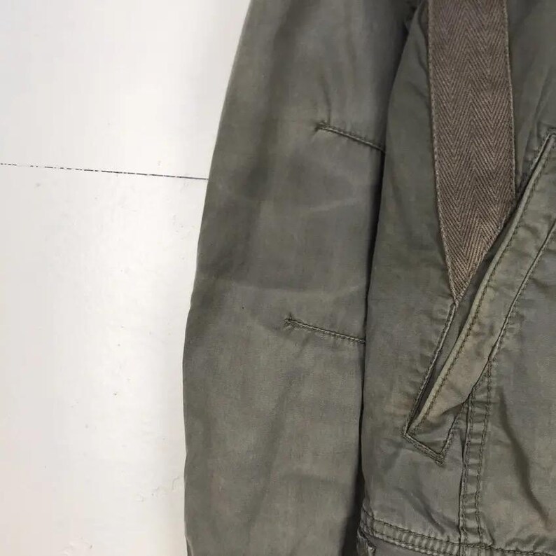 Puede incluir: Primer plano de una chaqueta verde oliva con un detalle de patr&oacute;n de espiga marr&oacute;n en el hombro. La chaqueta tiene un bolsillo y detalles de costura visibles. La tela parece ser un material duradero, posiblemente resistente al agua.